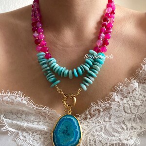 May include: A layered necklace with pink and turquoise beads and a large turquoise agate pendant with a gold setting. The necklace is on a model wearing a white lace top.