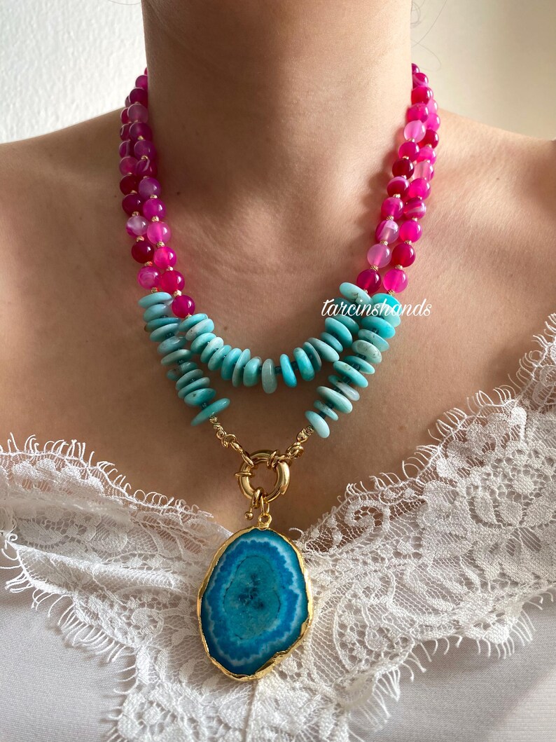 May include: A layered necklace with pink and turquoise beads and a large turquoise agate pendant with a gold setting. The necklace is on a model wearing a white lace top.