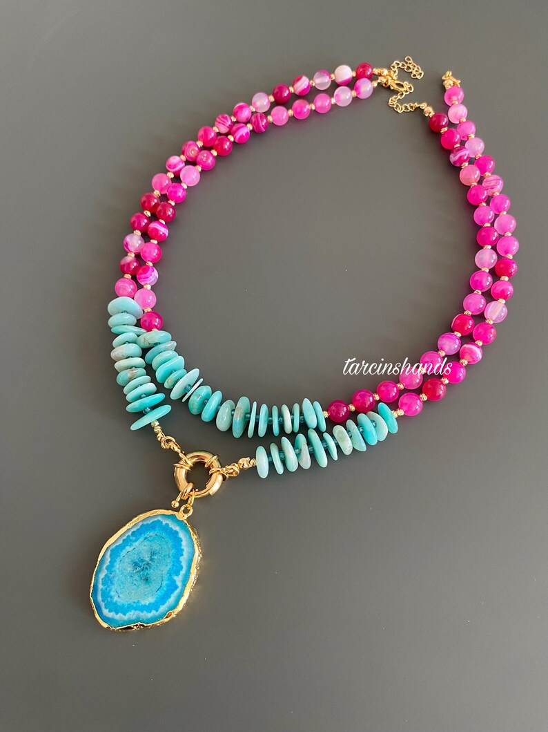 May include: A layered necklace with a large blue agate pendant. The top layer is made of pink and white agate beads, and the bottom layer is made of turquoise beads.
