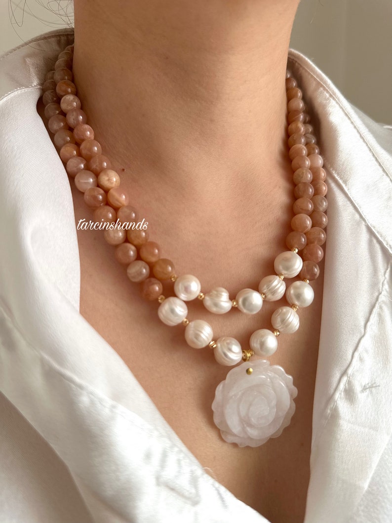 May include: A double-strand necklace with a rose-shaped pendant. The top strand is made of peach-colored sunstone beads, and the bottom strand is made of white pearls. The pendant is made of white jade and has a carved rose design.