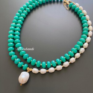 Puede incluir: Un collar de dos hilos con cuentas de color turquesa y perlas blancas. El collar tiene detalles dorados y un gran colgante de perla en forma de pera. El collar se muestra sobre una superficie gris oscuro.