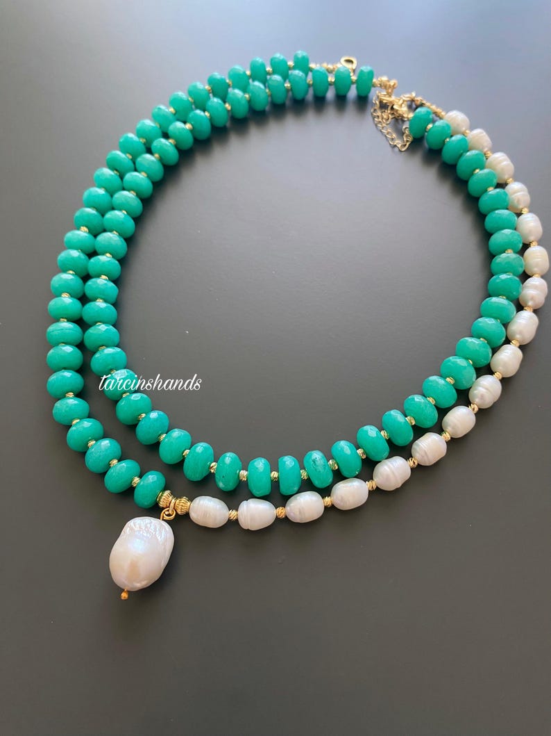 Puede incluir: Un collar de dos hilos con cuentas de color turquesa y perlas blancas. El collar tiene detalles dorados y un gran colgante de perla en forma de pera. El collar se muestra sobre una superficie gris oscuro.