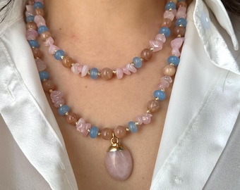Könnte beinhalten: Eine mehrreihige Halskette mit rosa, blauen und beigefarbenen Edelsteinperlen. Die Halskette hat einen goldfarbenen Verschluss und einen großen, ovalen rosa Anhänger. Die Halskette wird über einem weißen Hemd getragen.