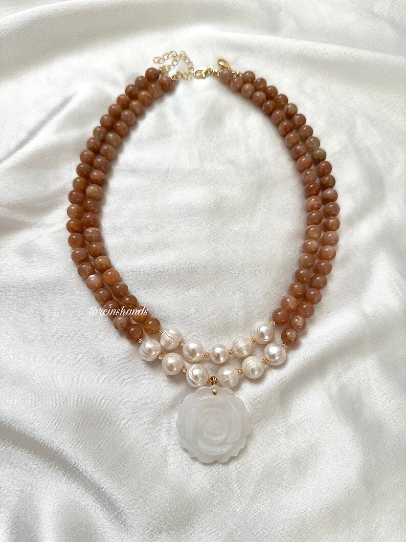 May include: A double-strand necklace with brown sunstone beads and white pearls. The necklace has a large white carved rose pendant.