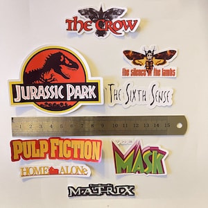 May include: A collection of movie-themed stickers featuring titles like "Jurassic Park," "Pulp Fiction," and "The Matrix." The stickers vary in shape and color, with bold text and graphic elements. A ruler is included for scale.