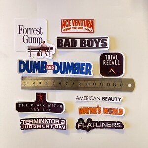 May include: A collection of movie-themed stickers, including "Forrest Gump," "Ace Ventura," "Bad Boys," "Dumb and Dumber," "Total Recall," "The Blair Witch Project," "American Beauty," "Wayne's World," "Terminator 2," and "Flatliners." A ruler shows measurements up to 6 inches.