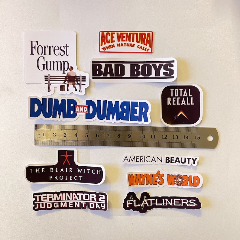 May include: A collection of movie-themed stickers, including "Forrest Gump," "Ace Ventura," "Bad Boys," "Dumb and Dumber," "Total Recall," "The Blair Witch Project," "American Beauty," "Wayne's World," "Terminator 2," and "Flatliners." A ruler shows measurements up to 6 inches.