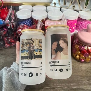 May include: Two frosted glass tumblers with lids and straws. Each tumbler features a photo and a music player design with song titles and artist names. The first tumbler has a photo of a couple in front of a Ferris wheel and the song "How To Love" by Lil Wayne. The second tumbler has a photo of two women and the song "Grateful" by DJ Khaled.