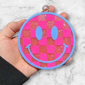 Puede incluir: Un adorno de cara sonriente rosa y azul con un patr&oacute;n a cuadros. El adorno est&aacute; hecho de resina y tiene un acabado brillante.