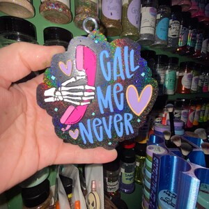 May include: A glittery, black keychain with the words "CALL ME NEVER" in blue, pink, and purple. The design includes a skeleton hand holding a pink phone and two purple hearts. The keychain has a rainbow glitter border.