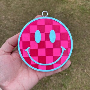 Puede incluir: Un amuleto de cara sonriente rosa y azul con un patr&oacute;n a cuadros. El amuleto est&aacute; hecho de pl&aacute;stico y tiene un lazo para colgar.