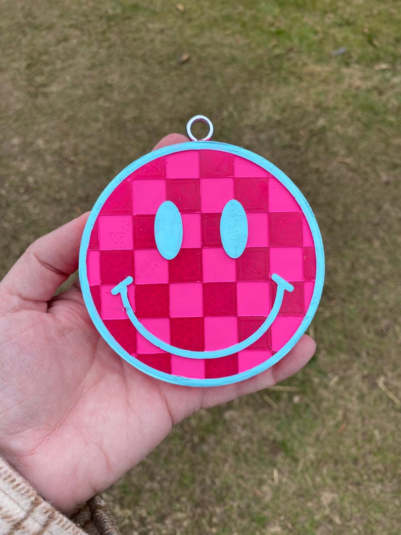 Puede incluir: Un amuleto de cara sonriente rosa y azul con un patr&oacute;n a cuadros. El amuleto est&aacute; hecho de pl&aacute;stico y tiene un lazo para colgar.