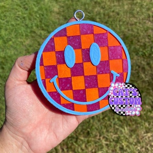 Puede incluir: Un adorno de cara sonriente a cuadros rosa y naranja con un contorno azul. El adorno tiene un lazo plateado para colgar. El adorno est&aacute; hecho de resina y tiene un acabado brillante.