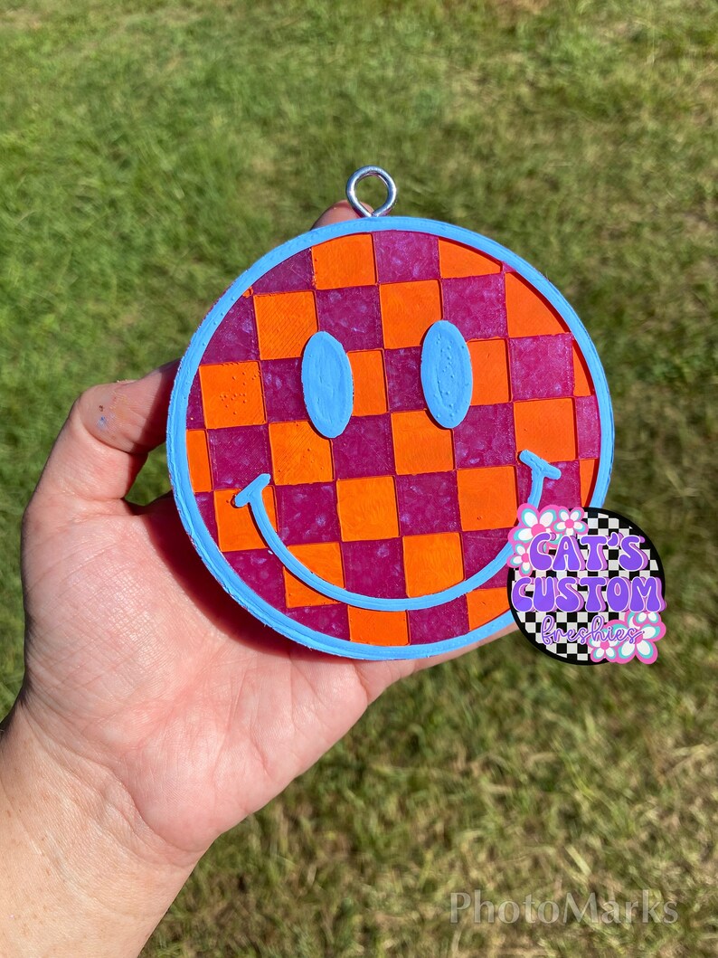 Puede incluir: Un adorno de cara sonriente a cuadros rosa y naranja con un contorno azul. El adorno tiene un lazo plateado para colgar. El adorno est&aacute; hecho de resina y tiene un acabado brillante.