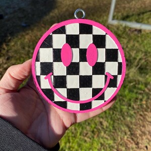 Puede incluir: Un adorno de cara sonriente a cuadros blanco y negro con ojos rosas y contorno rosa. El adorno tiene un lazo plateado para colgar.