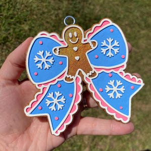 May include: A Christmas ornament shaped like a bow with a gingerbread man in the center. The bow has blue sections with white snowflakes and pink trim. The gingerbread man is gold with a smiling face.