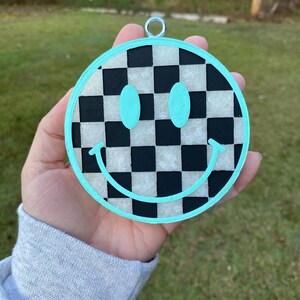 Puede incluir: Un adorno de cara sonriente azul turquesa con un patr&oacute;n de cuadros negros y blancos. El adorno tiene un lazo plateado para colgar.