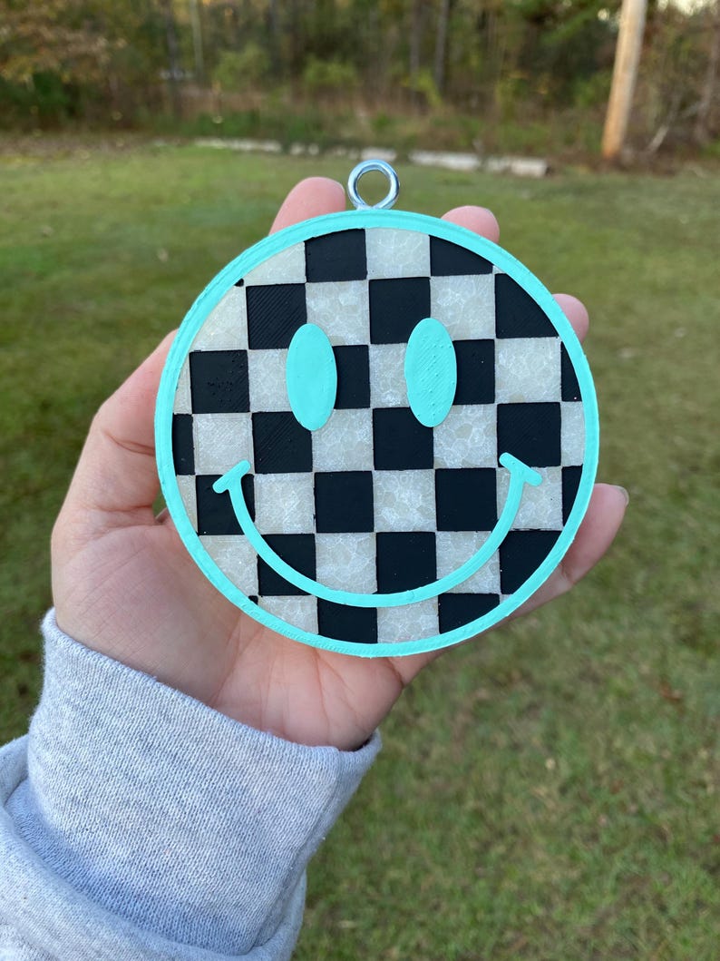 Puede incluir: Un adorno de cara sonriente azul turquesa con un patr&oacute;n de cuadros negros y blancos. El adorno tiene un lazo plateado para colgar.
