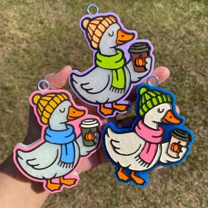 May include: Three whimsical duck ornaments, each holding a coffee cup with a pumpkin design. The ducks wear colorful hats and scarves in shades of orange, green, blue, and pink. Each ornament has a silver loop for hanging.