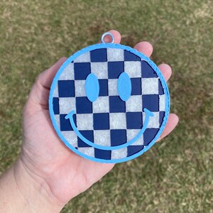 Puede incluir: Un adorno de cara sonriente a cuadros azules y blancos con un contorno azul claro. El adorno tiene un lazo plateado para colgar.