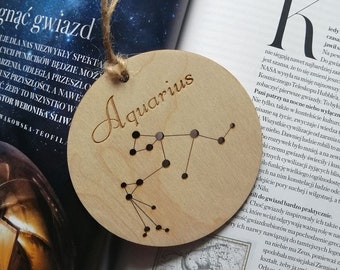 May include: A wooden circle ornament with the constellation Aquarius engraved in black. The word "Aquarius" is written in a cursive font above the constellation.