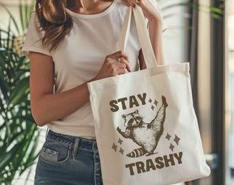May include: White canvas tote bag with brown text that reads "Stay Trashy" and a cartoon raccoon wearing a sparkly headband.