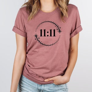 May include: A mauve coloured t-shirt with a black circle design featuring the number 11:11 and small stars around the circle.