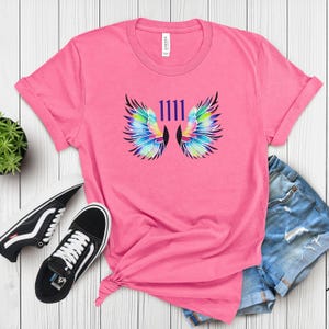 May include: Pink t-shirt featuring a colourful graphic of angel wings and the number 1111 in purple.