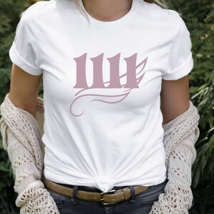 May include: White t-shirt featuring a pink graphic of the Roman numeral 'III' with a stylised wing design.