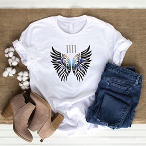 May include: White t-shirt with a graphic of angel wings and the number "1111" in grey. The wings are black with blue and purple details. The shirt is styled with blue jeans and tan ankle boots.