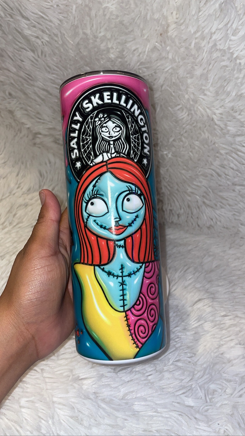 May include: A white stainless steel tumbler with a colourful design featuring Sally from The Nightmare Before Christmas. The design includes a pink background with the text "Sally Skellington" in a black circle. Sally is depicted with her signature red hair, blue skin, and patchwork clothing.