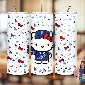 May include: A white stainless steel tumbler with a Hello Kitty design wearing a Los Angeles Dodgers baseball cap and jersey. The tumbler has a white background with a repeating pattern of blue baseballs, red bows, and the letters "LA" in blue.