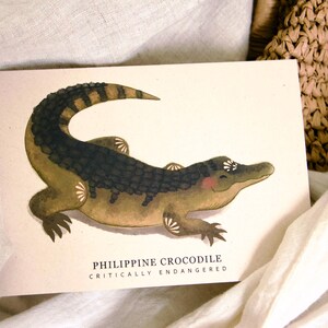 May include: A watercolor illustration of a Philippine crocodile on a cream-colored card. The crocodile is depicted in shades of green, brown, and black, with a smiling expression. Text at the bottom reads "Philippine Crocodile Critically Endangered."