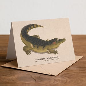 May include: A greeting card featuring an illustration of a Philippine crocodile in shades of green, brown, and black. The card is on a kraft paper envelope, with the text "PHILIPPINE CROCODILE CRITICALLY ENDANGERED". The card is on a wooden surface.