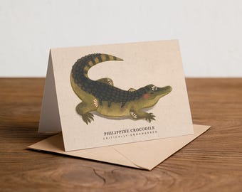 Może przedstawiać: Kartka okolicznościowa z ilustracją krokodyla filipińskiego w odcieniach zieleni, brązu i czerni. Karta leży na kopercie z papieru kraft. Tekst brzmi "PHILIPPINE CROCODILE CRITICALLY ENDANGERED". Na drewnianej powierzchni.