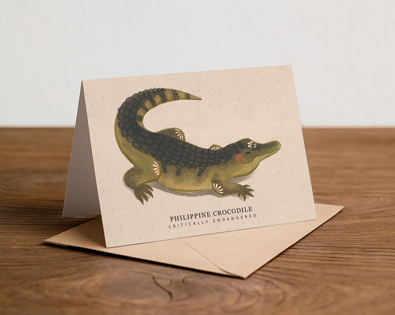 May include: A greeting card featuring an illustration of a Philippine crocodile in shades of green, brown, and black. The card is on a kraft paper envelope, with the text "PHILIPPINE CROCODILE CRITICALLY ENDANGERED". The card is on a wooden surface.