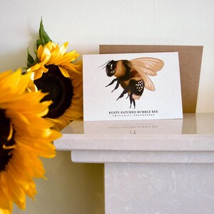以下が含まれることがあります： 「Rusty-Patched Bumble Bee Critically Endangered」のテキストが入った、錆びた斑点のあるマルハナバチの詳細なイラストが描かれたグリーティングカード。 カードには茶色の封筒と明るい黄色のヒマワリが添えられています。