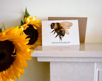 Może przedstawiać: Kartka okolicznościowa z szczegółową ilustracją trzmiela rdzawego z napisem "Rusty-Patched Bumble Bee Critically Endangered". Karta jest w towarzystwie brązowej koperty i jasnych żółtych słoneczników.