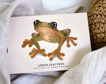 Könnte beinhalten: Aquarell-Illustration eines Lemur-Blattfrosches, vom Aussterben bedroht, auf einer cremefarbenen Karte. Der Frosch ist in Grün-, Braun- und Beigetönen mit großen schwarzen Augen dargestellt. Die Karte befindet sich vor einem Hintergrund aus weißem Stoff und einem geflochtenen Korb.
