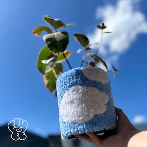 May include: A blue and white planter with a cloud design. The planter is made of a fluffy material and is being held by a hand.