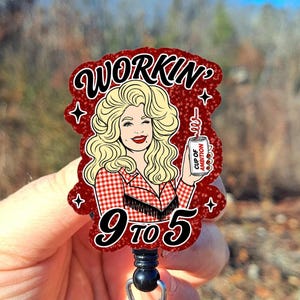 May include: A badge reel featuring a glittery red background with a cartoon illustration of a blonde person in a red and white checkered shirt. The text reads Workin' 9 to 5 and Cup of Ambition on a silver can held by the illustrated figure.