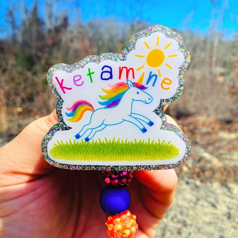 May include: A glittery badge reel featuring a white unicorn with a rainbow mane and tail, running on green grass under a yellow sun. The word ketamine is printed in colourful letters above the unicorn. The reel has a purple and orange beaded attachment.