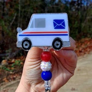May include: A white USPS mail truck badge reel with red and blue stripes, a blue envelope graphic, and glitter accents. The badge reel has red, white, and blue beads.