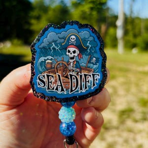 May include: A pirate-themed badge reel with a skull and crossbones design. The badge reel features a pirate skeleton steering a ship in a storm, with the words "SEA DIFF" in white. The edges are outlined with black glitter and the reel has blue and turquoise beads.