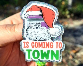 May include: A festive badge with a Santa hat design, featuring a jar labeled "Sani-Cloth" and the text "IS COMING TO TOWN" in red and green. The badge has a glittery border and a gold bead.