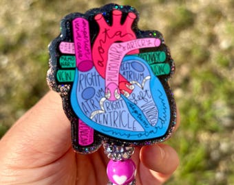 May include: A heart-shaped badge reel with a detailed anatomical illustration of the heart. The design features a blue and pink heart with labeled parts, surrounded by a black glitter border. The reel has a beaded accent.