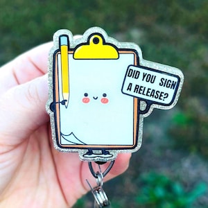 May include: A cute badge reel featuring a smiling clipboard character with a yellow pencil. The clipboard has the text "DID YOU SIGN A RELEASE?" in a speech bubble. The badge has a glittery border and a retractable cord.