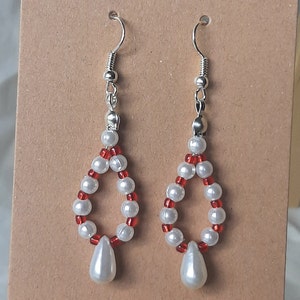 May include: A pair of dangle earrings with a teardrop-shaped pearl hanging from a silver hook. The earrings are made with white and red beads.