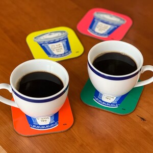 May include: Two white coffee mugs with blue trim, filled with black coffee, sitting on colorful coasters with the text "I love you" in a blue font. The coasters are orange, green, and red.