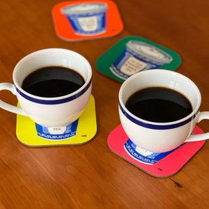 May include: Two white coffee mugs with black coffee on yellow and red coasters with a blue and white design. The coasters have the text "YOU" and "ASG" on them.